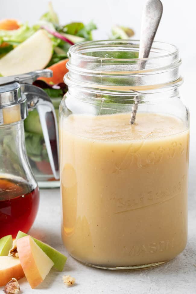 A clear glass mason jar filled with maple vinaigrette. A salad with apple slices sits next to the mason jar.