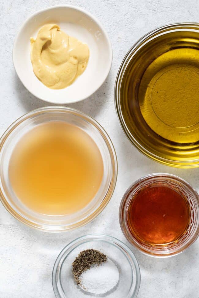 Clear glass measuring bowls filled with olive oil, apple cider vinegar, pure maple syrup and Dijon mustard.