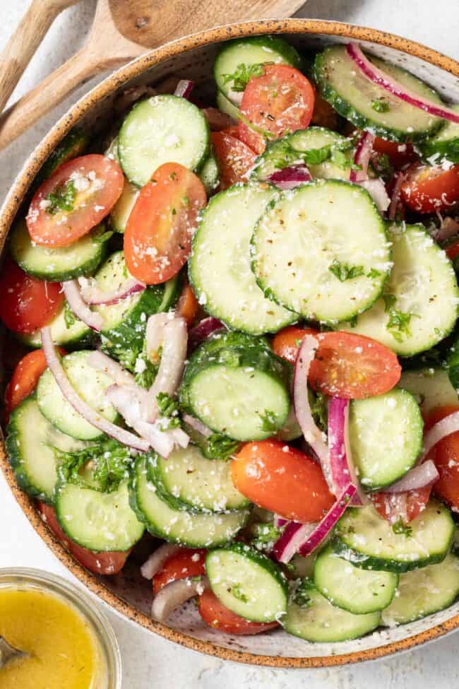 A tan colored bowl filled with Cucumber Tomato Salad. Wooden salad spoons rest in the bowl.