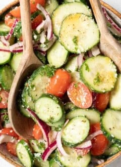A tan colored bowl filled with cucumber tomato salad drizzled with red wine vinaigrette. Wooden salad servers rest in the bowl.
