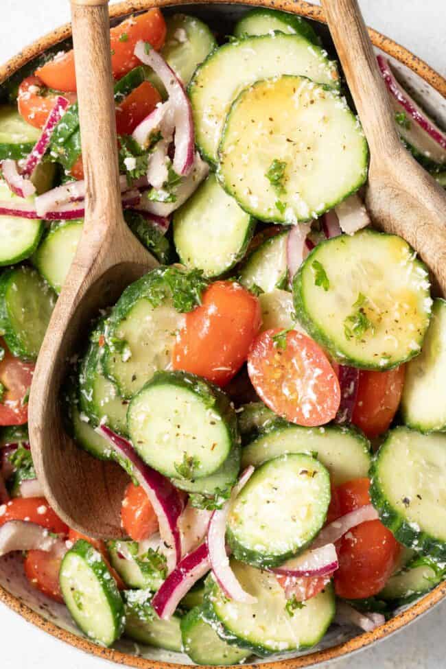 A tan colored bowl filled with Cucumber Tomato Salad. Wooden salad spoons rest in the bowl.