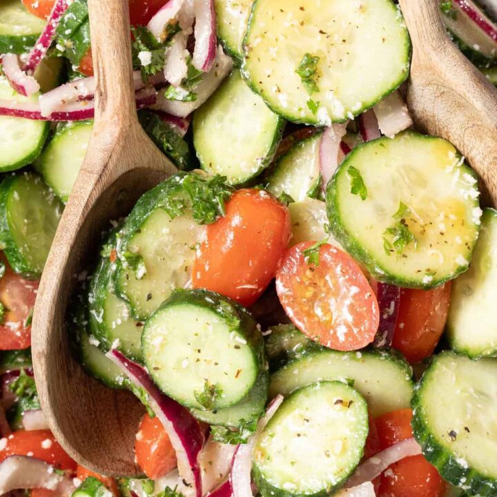 A tan colored bowl filled with cucumber tomato salad drizzled with red wine vinaigrette. Wooden salad servers rest in the bowl.