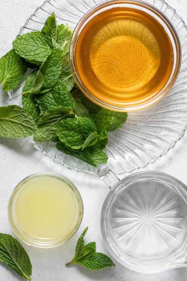 Three glass bowls filled with lemon juice, honey and water. The honey sits on a glass plate scattered with fresh herbs.