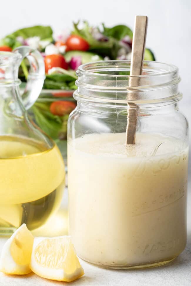 A clear mason jar filled with lemon olive oil dressing. A clear glass bowl filled with green salad sits next to the mason jar.