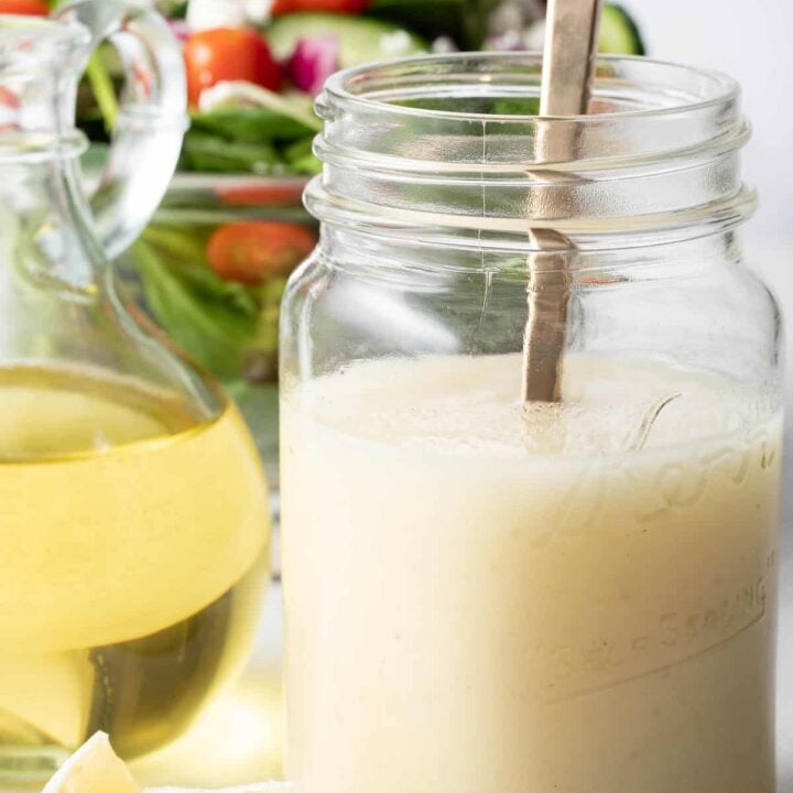 A clear mason jar filled with lemon olive oil dressing. A clear glass bowl filled with green salad sits next to the mason jar.