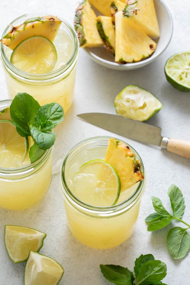 Three mason jars are filled with pineapple and mint green tea. Sprigs of mint are scattered next to the jars and a small bowl with pieces of pineapple sits near the jars.