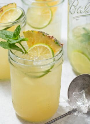 Four glass mason jars filled with Iced Pineapple Mint Green Tea. One of the glasses is garnished with Mint, pineapple wedges and slices of lime.