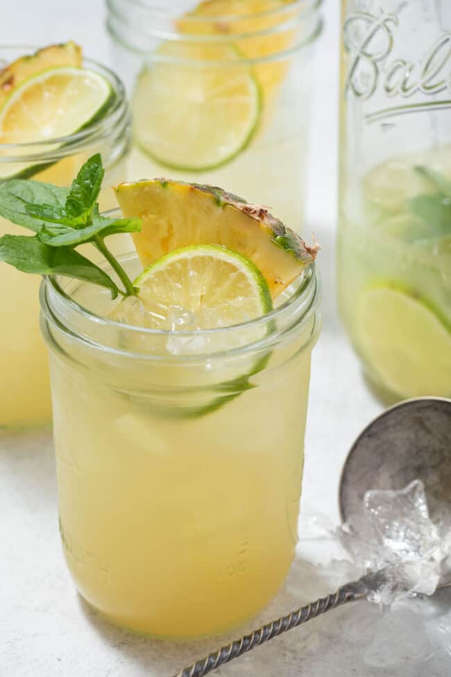 Four glass mason jars filled with Iced Pineapple Mint Green Tea. One of the glasses is garnished with Mint, pineapple wedges and slices of lime.