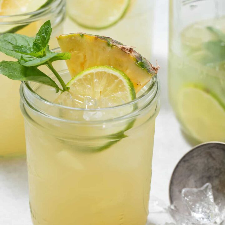 Four glass mason jars filled with Iced Pineapple Mint Green Tea. One of the glasses is garnished with Mint, pineapple wedges and slices of lime.