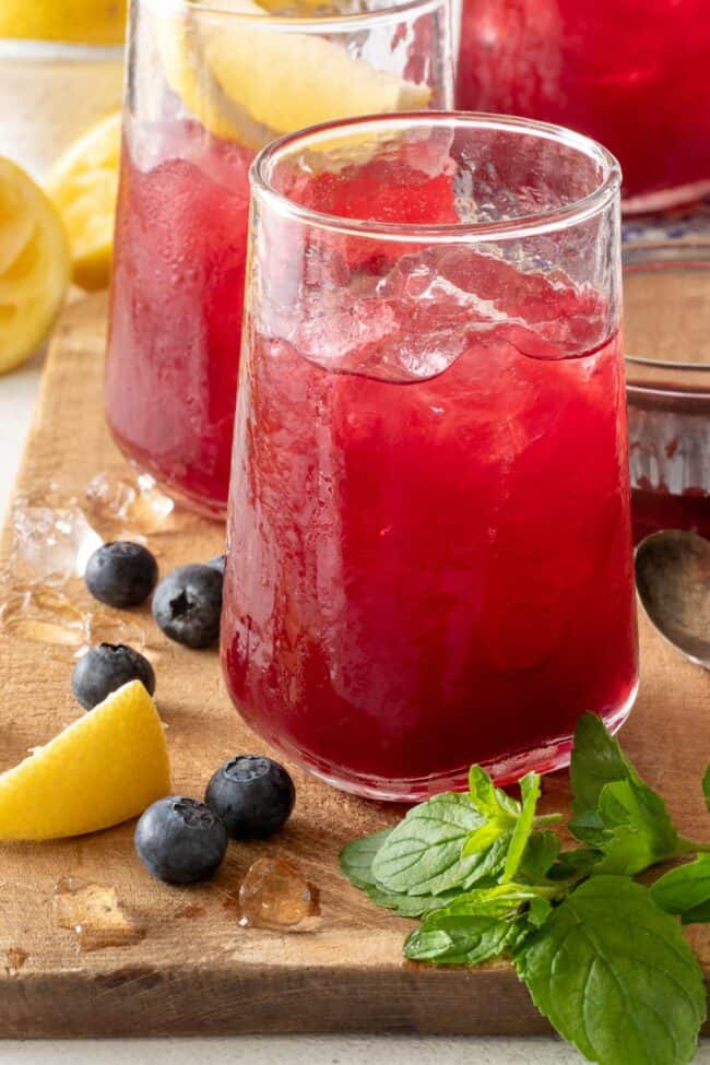 Three clear drinking glasses filled with Blueberry Lemonade. The glasses are sitting on a wooden cutting board with blueberries and lemon wedges.