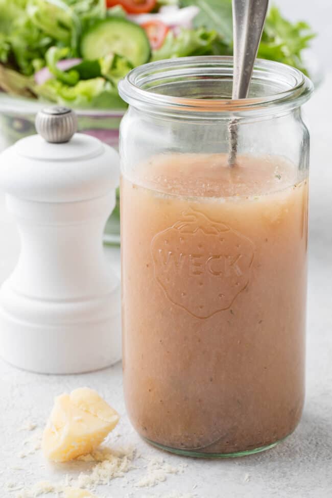 A clear mason jar filled with homemade red wine vinaigrette sits next to a clear glass bowl filled with salad.