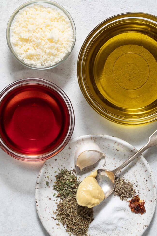 Three small glass mixing bowls filled with red wine vinegar, olive oil and grated parmesan cheese. A small plate filled with spices sits next to the bowls.