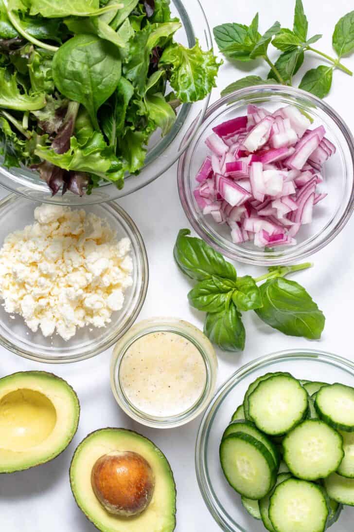 Four glass mixing bowls filled with cucumber slices, chopped onion, Feta cheese and baby lettuce.
