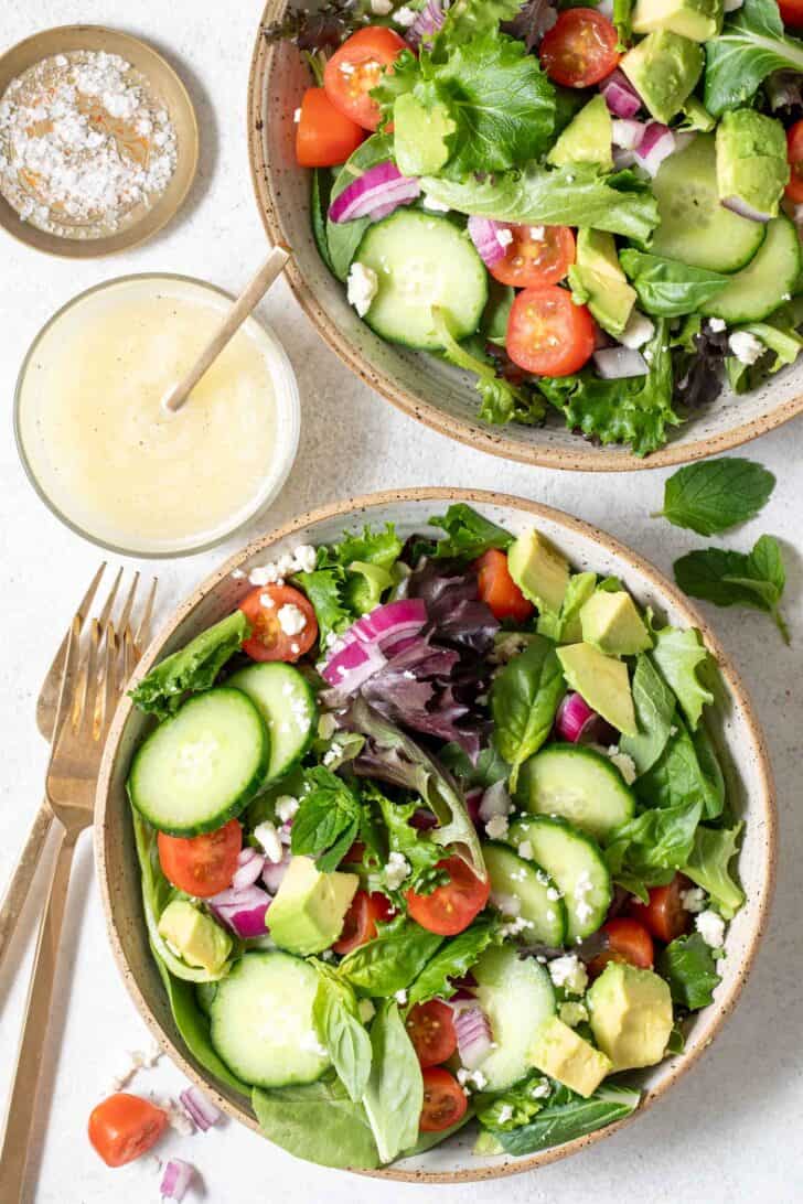 Two tan colored bowls filled with mesclun salad. A small glass bowl is filled with dressing. Two gold forks sit next to the bowls.