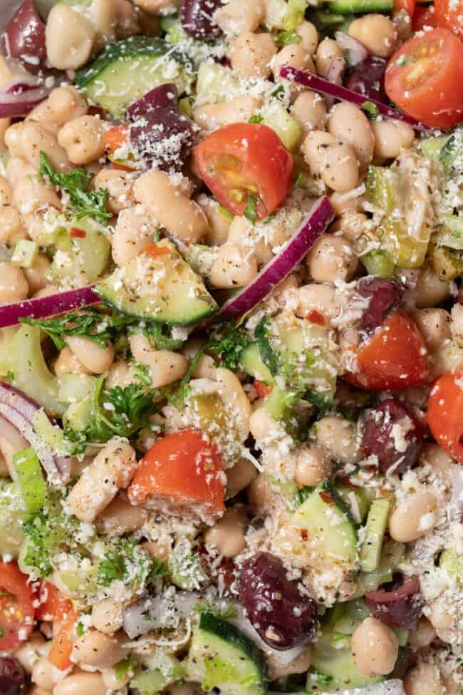 A close up photo of white bean salad with chopped vegetables and Parmesan cheese tossed in a red wine vinaigrette. A serving spoon rests in the salad.