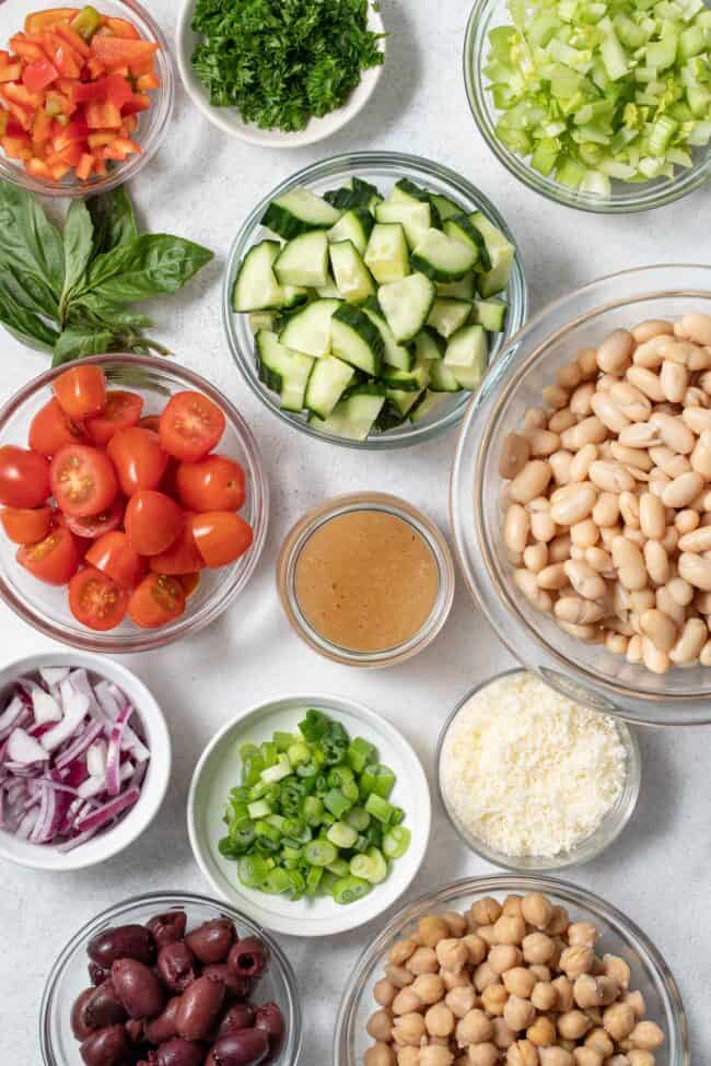 Glass measuring bowls filled with legumes, vegetables, olives, herbs and grated cheese.