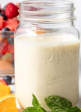 A clear glass mason jar filled with Yogurt Fruit Salad Dressing. A clear glass bowl filled with fruit salad sits next to the jar.