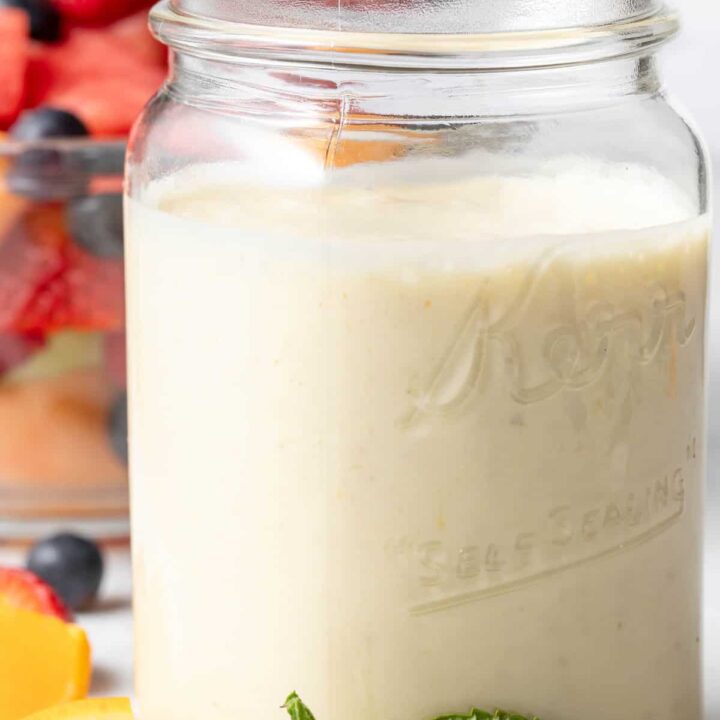 A clear glass mason jar filled with Yogurt Fruit Salad Dressing. A clear glass bowl filled with fruit salad sits next to the jar.