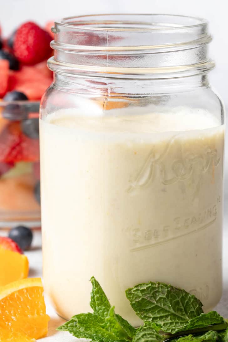 A clear mason jar filled with creamy fruit salad dressing. A clear glass bowl filled with fruit salad sits next to the jar.