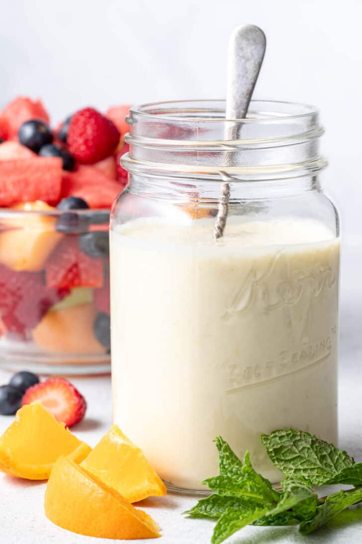 A clear mason jar filled with Greek Yogurt Fruit Salad Dressing. A clear glass bowl filled with fresh fruit sits next to the mason jar.