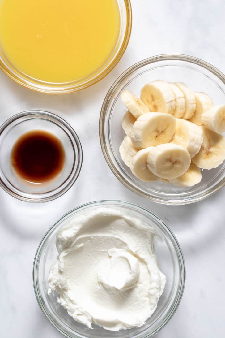 Four clear glass mixing bowls filled with orange juice, sliced banana, vanilla extract and dairy product.