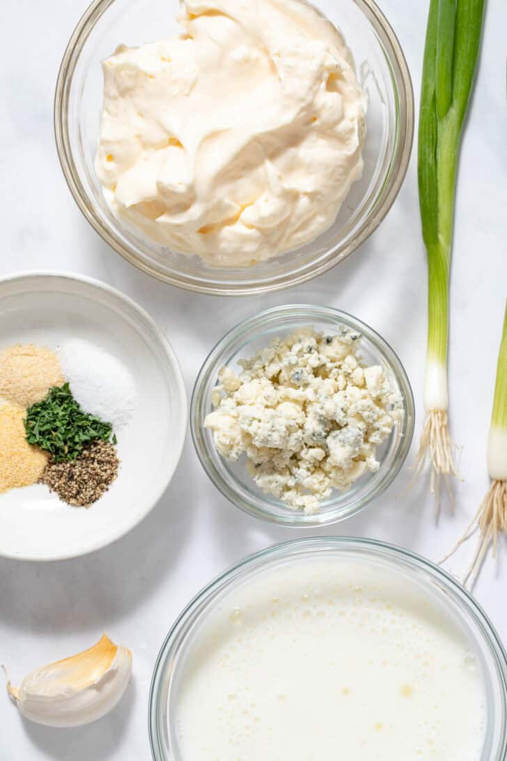 Small clear glass mixing bowls filled with mayonnaise, buttermilk, Gorgonzola cheese and seasoning.