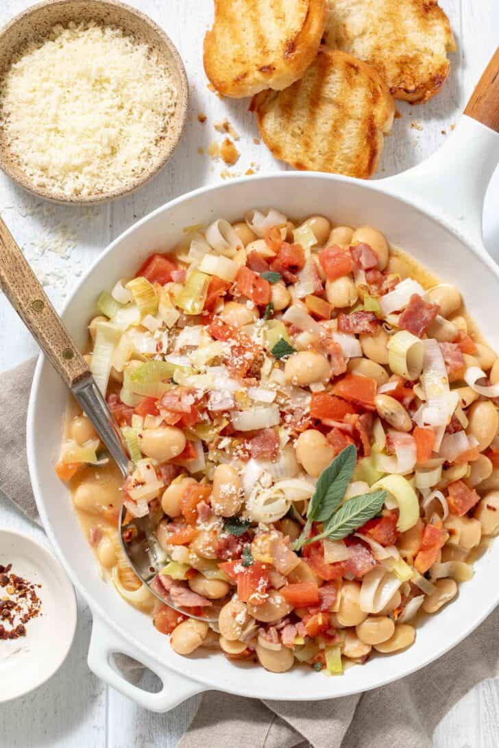 A white skillet filled with legumes cooked with bacon, tomatoes, garlic and leeks.