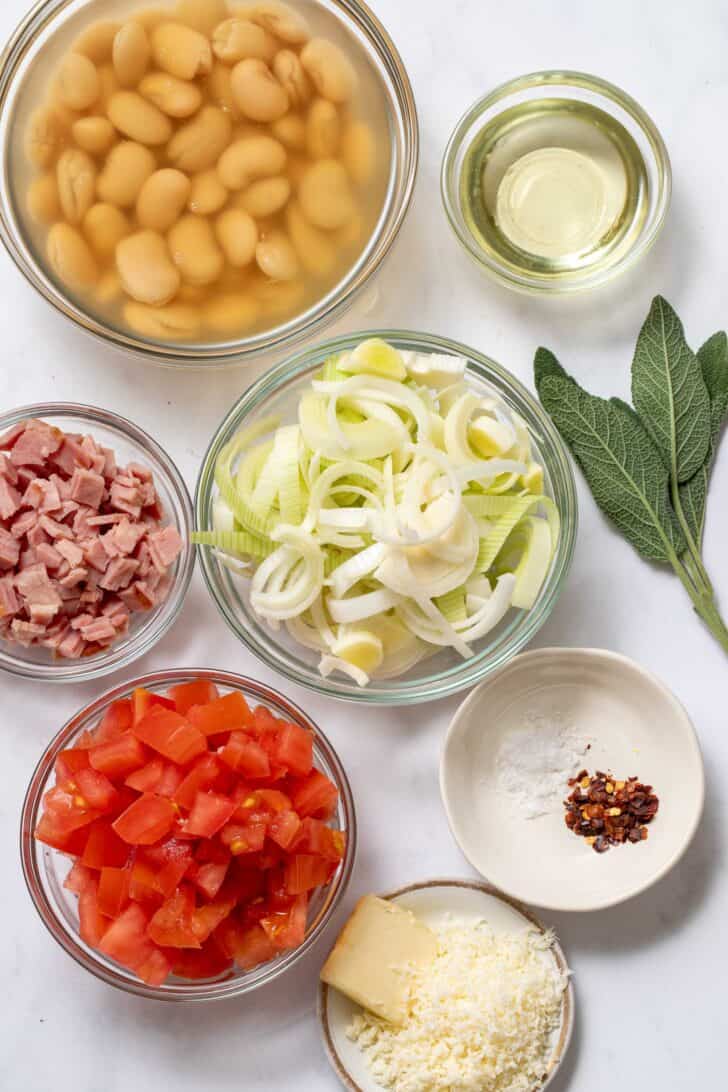 Four clear glass bowls filled with canned butter beans, sliced leeks, chopped turkey bacon and chopped tomatoes.