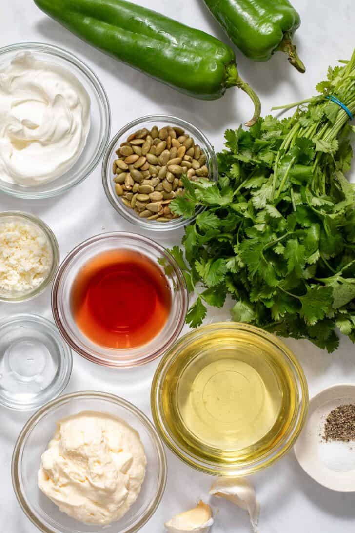 Small clear glass mixing bowls filled with mayonnaise, sour cream, olive oil, vinegar, and cheese. A bunch of cilantro and two peppers sit next to the bowls.