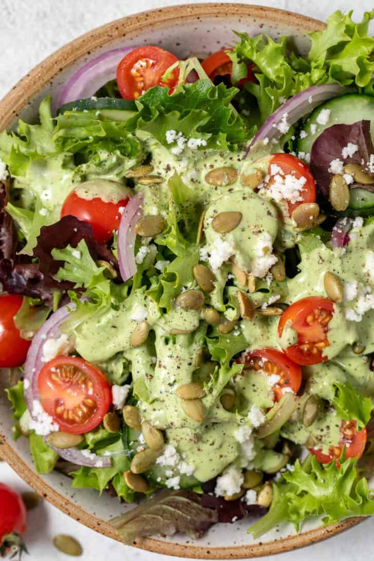 A tan colored bowl filled with mixed greens, cherry tomatoes, red onions and pepitas tossed in a Creamy Cilantro Salad Dressing.