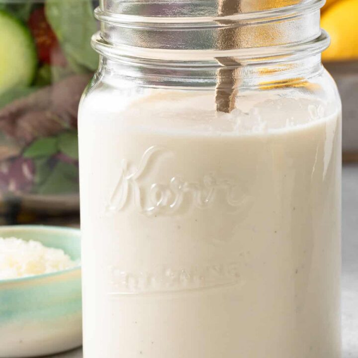 A clear glass mason jar filled with Creamy Lemon Salad Dressing. A clear glass bowl filled with mixed greens sits behind the jar.