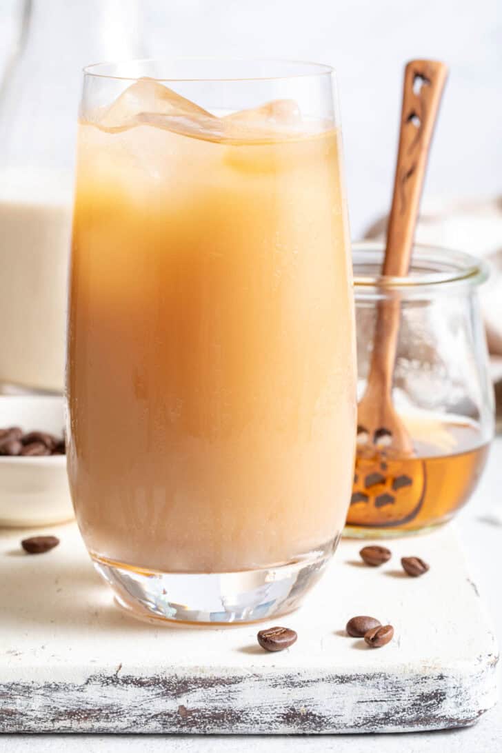 A clear glass filled with a cold drink. A bottle of oat milk and a small jar. of honey sits next to the glass.