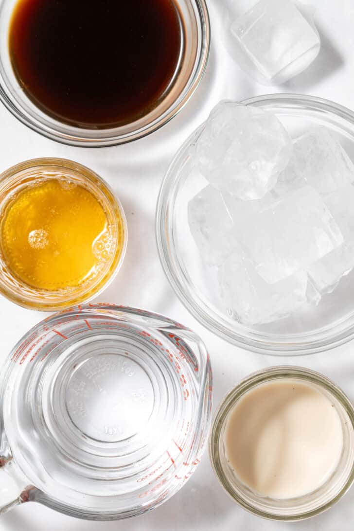 Four small clear glass bowls filled with ice, water, oat milk and honey.