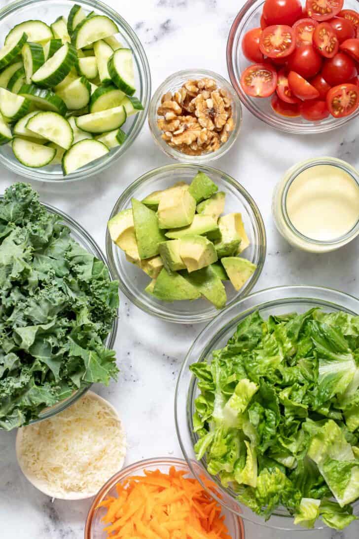 Clear glass bowls filled with chopped lettuce, avocado, grated carrots, sliced cucumber, cherry tomatoes and waluts.