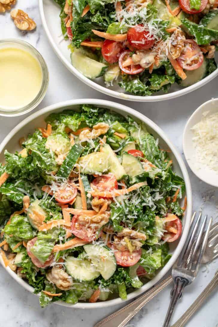 Two white bowls filled with lemon kale parmesan salad. Silver colored forks sit next to the bowls.