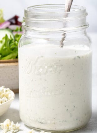 A clear mason jar filled with Blue Cheese Ranch Dressing. A tan bowl filled with salad sits behind the jar.