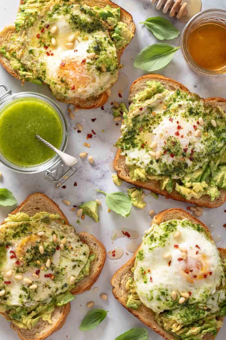Four pieces of Pesto Eggs Avocado Toast. A glass jar filled with pesto sits next to the toast.