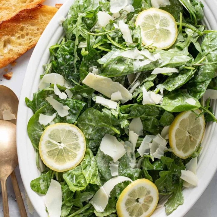A white oval platter filled with Lemon Arugula Salad tossed with Parmesan Shavings.