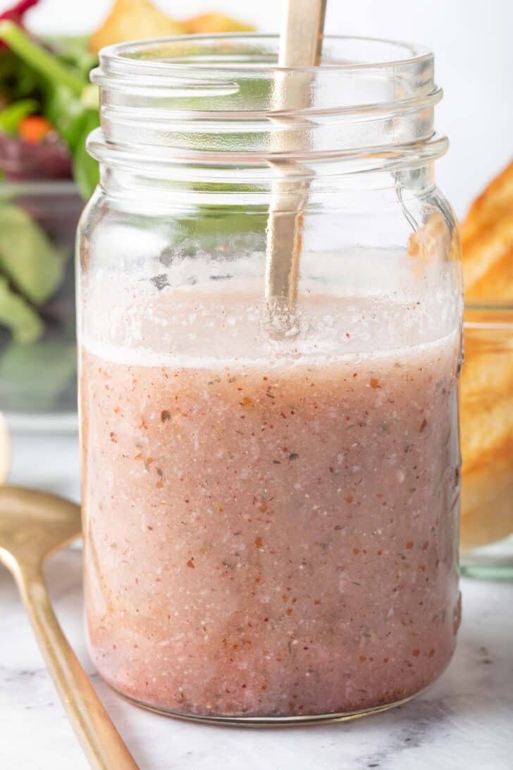 A clear glass mason jar filled with Lemon Sumac Salad Dressing. A clear glass bowl filled with salad sits next to the jar.