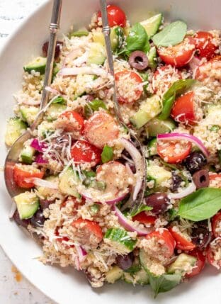 A white bowl filled with Tomato Basil Quinoa Salad. Silver colored salad servers rest in the bowl.