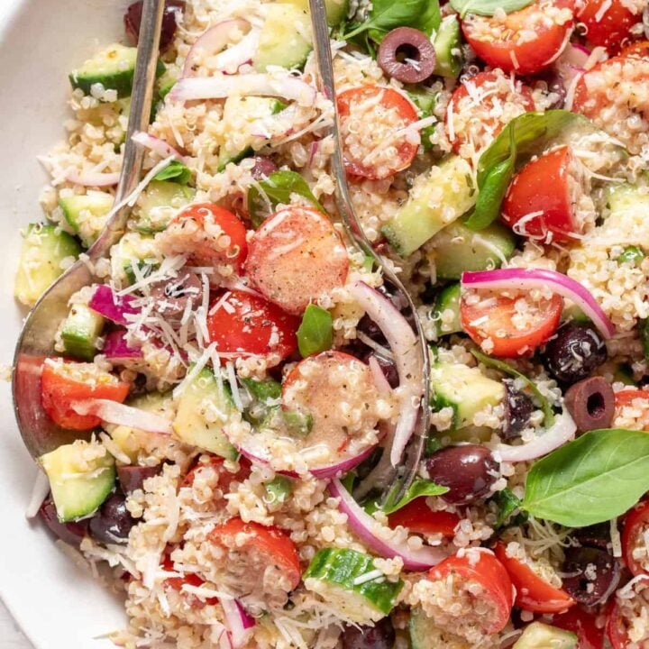A white bowl filled with Tomato Basil Quinoa Salad. Silver colored salad servers rest in the bowl.