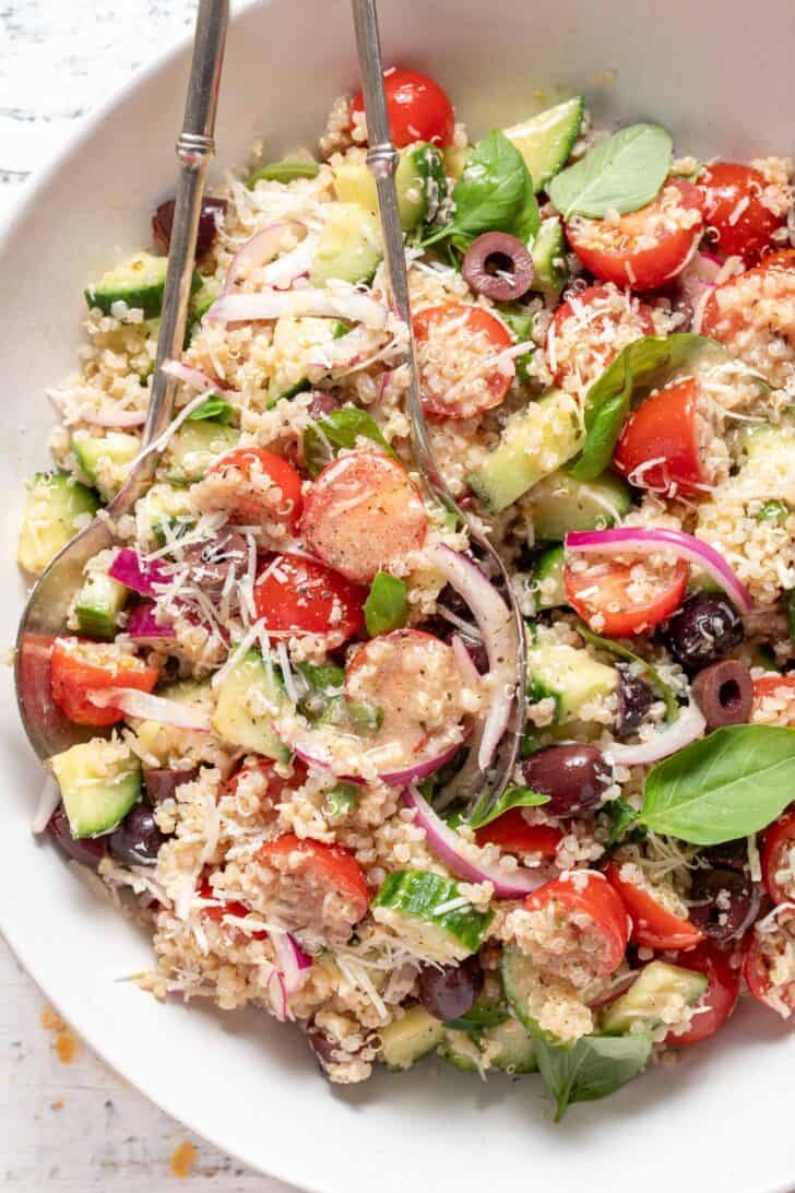 A white bowl filled with Tomato Basil Quinoa Salad. Silver colored salad servers rest in the bowl.