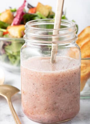 A clear glass mason jar filled with Sumac Salad Dressing. A clear glass bowl filled with salad sits next to the jar.