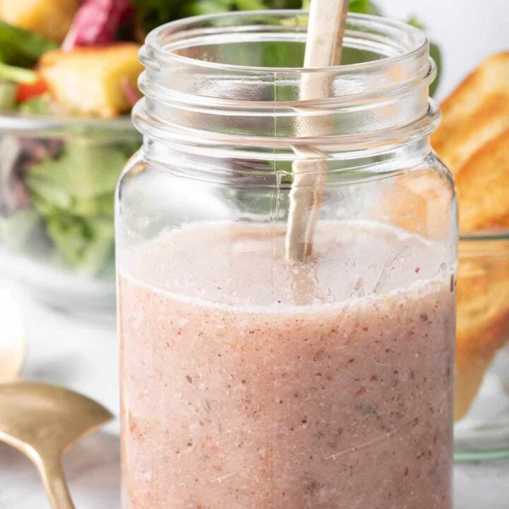A clear glass mason jar filled with Sumac Salad Dressing. A clear glass bowl filled with salad sits next to the jar.