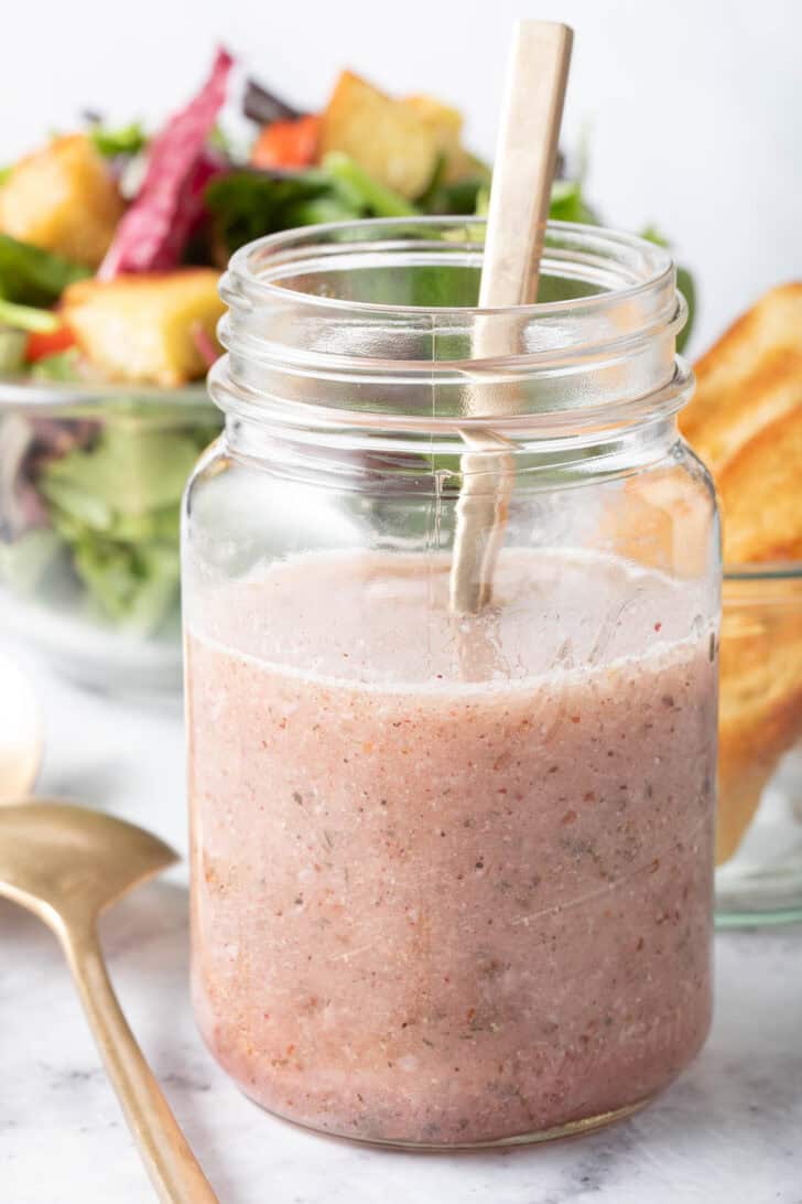 A clear glass mason jar filled with Sumac Salad Dressing. A clear glass bowl filled with salad sits next to the jar.