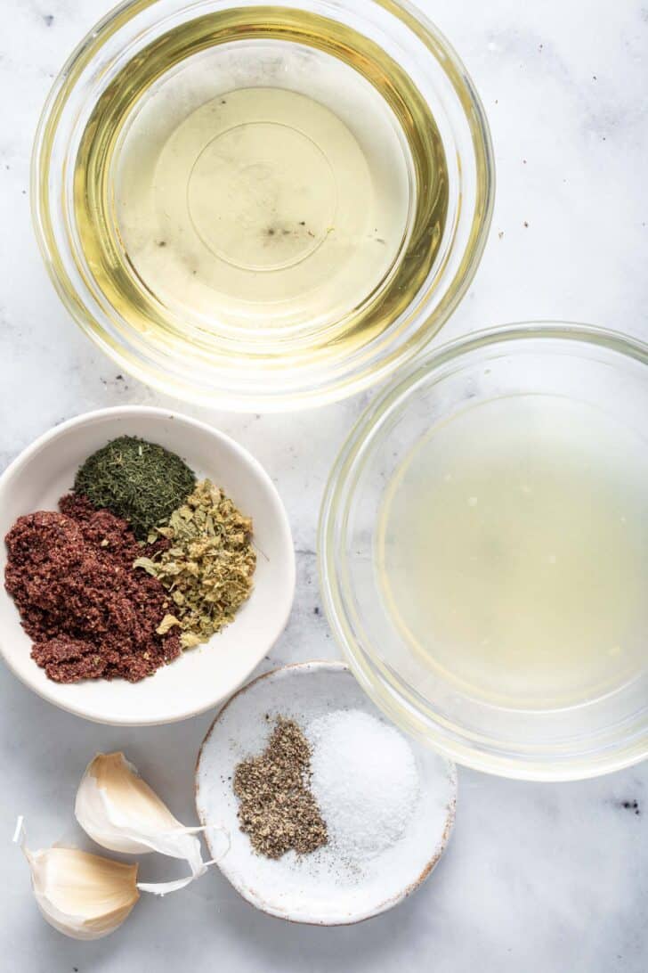 Two clear glass bowls filled with olive oil and lemon juice. A small white bowl filled with spices sits next to the glass bowls. For Sumac Salad Dressing.