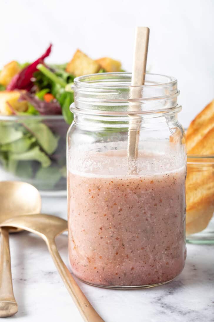 A clear glass mason jar filled with a pink vinaigrette. A clear glass bowl filled with salad sits next to the jar.