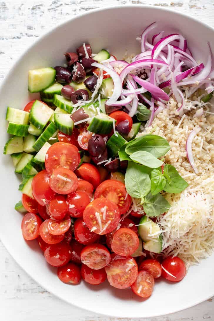 A white bowl filled with cucumber, olives, onion, herbs and a grain.