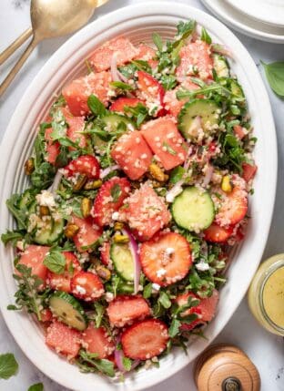 A white oval platter filled with watermelon arugula salad with feta cheese. Gold colored salad tongs sit next to the platter.