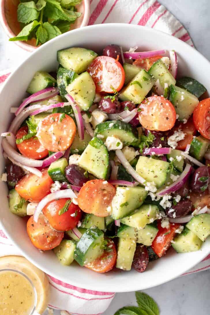 A white bowl filled with Greek Cucumber Salad tossed in dressing. A small glass filled with dressing sits next to the bowl.